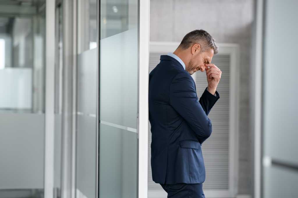 Tired business man suffering from headache at work. Mid man entrepreneur feeling stressed while pressing eye standing and leaning at glass wall in office. Depressed businessman suffering from migraine